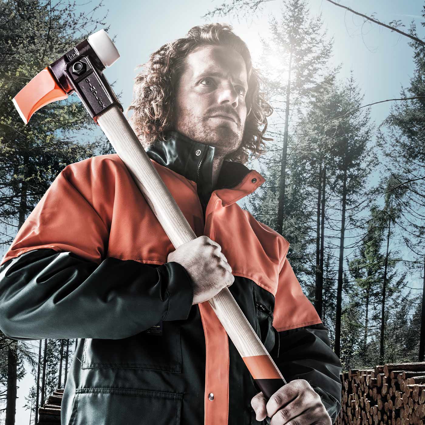 Homme en costume de travail orange et noir tenant la hache Simplex 900 sur son épaule, forêt et troncs d’arbres empilés en arrière-plan