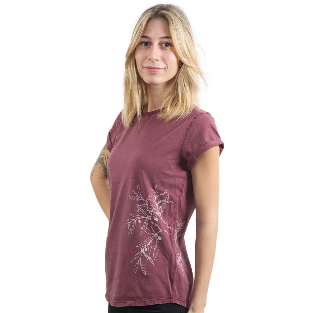 Une femme porte un T-shirt bordeaux « Rameau d’olivier » avec un motif de branche d’olivier blanc sur le côté et a les mains derrière le dos.