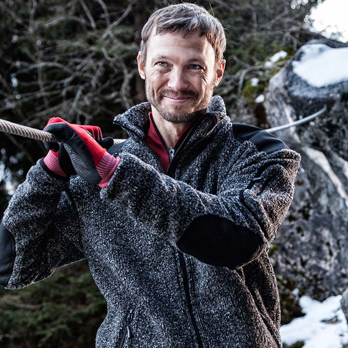 Homme portant une veste grizzly, un cardigan gris avec une bordure noire sur les épaules et les coudes, un maillot de corps rouge et des gants rouges, une forêt et des rochers avec de la neige en arrière-plan