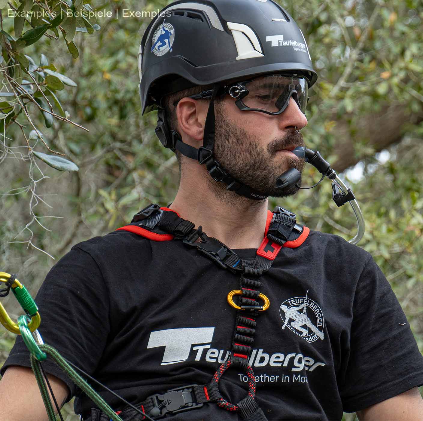 Homme portant un harnais d’escalade upMOTION SRT, un casque noir et des lunettes, tenant un tube d’abreuvement dans sa bouche, des arbres et des feuilles en arrière-plan