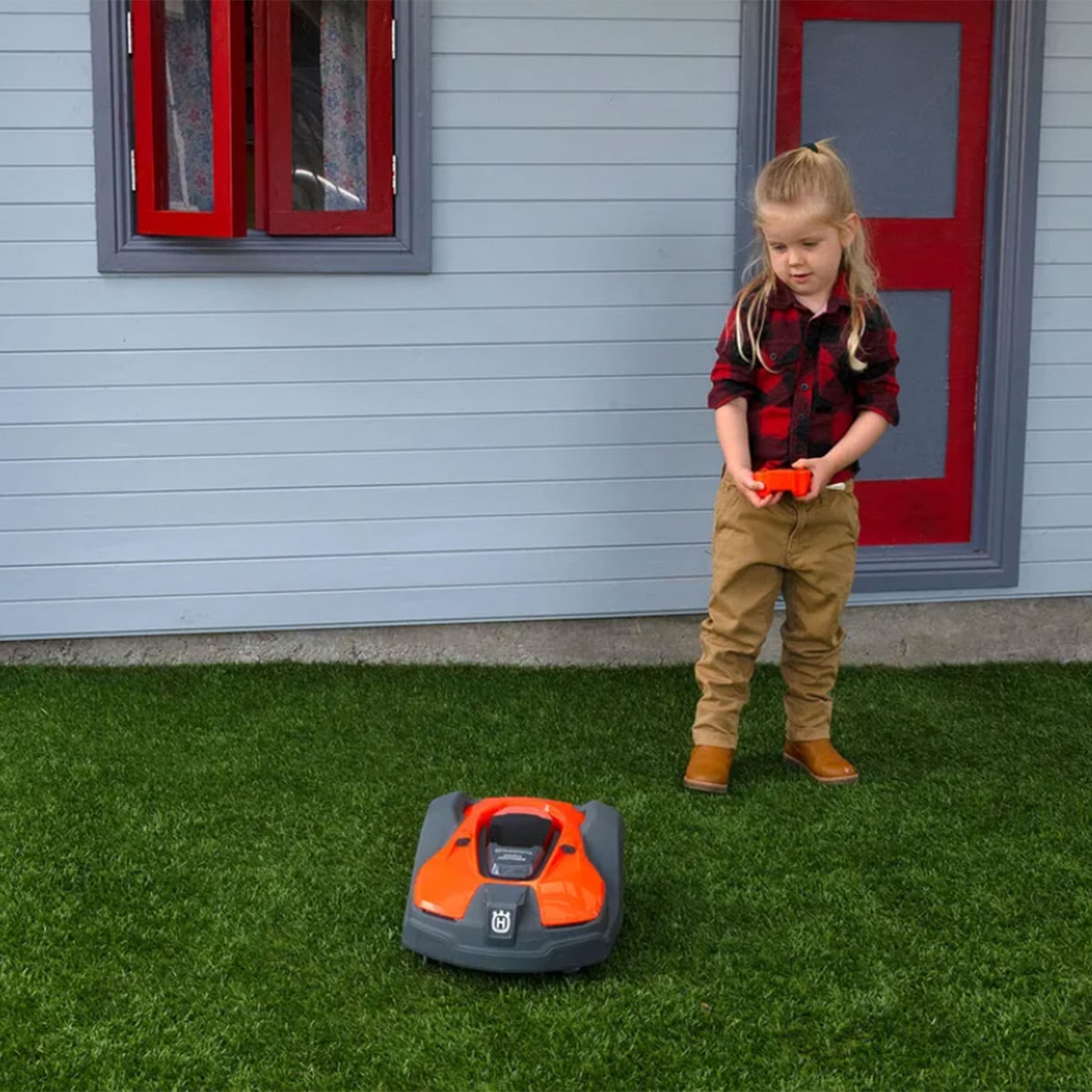 Un enfant vêtu d’une chemise à carreaux rouges, d’un pantalon beige et de bottes marron contrôle l’Automower orange et noir pour enfants de Husqvarna sur une pelouse verte devant une maison avec une fenêtre et un cadre de porte rouges