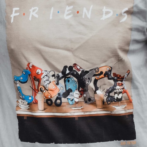 Gros plan de l’imprimé du T-shirt avec les mots « Friends » et des pièces mécaniques détaillées de différentes couleurs sur une table, avec le lettrage « ARB FICTION » visible en bas à droite