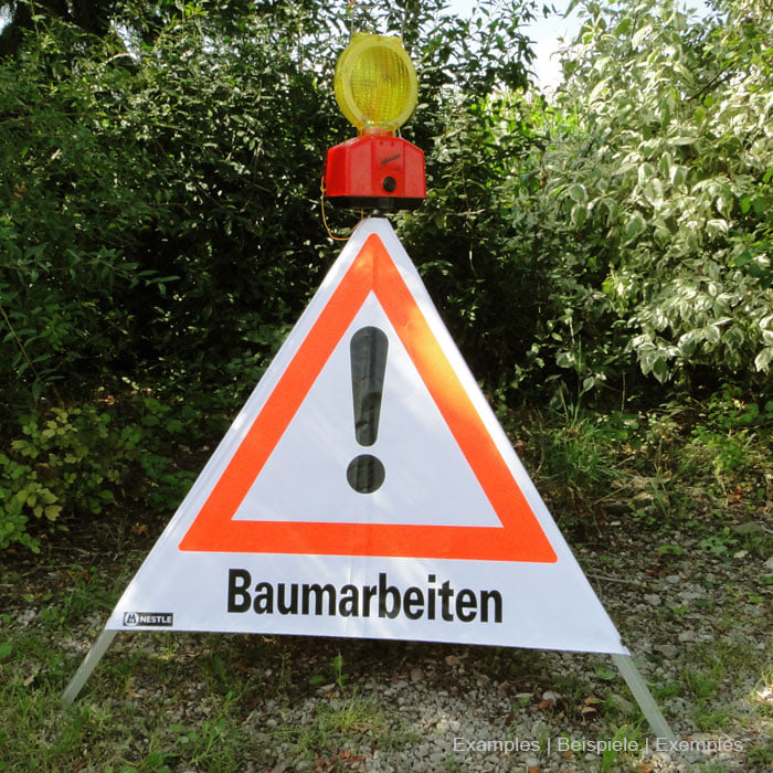 Triangle pliant d’avertissement avec l’impression « Travail des arbres » et un point d’exclamation dans un triangle orange, avec un voyant d’avertissement jaune et un boîtier rouge fixé au sommet, debout sur de l’herbe et du gravier