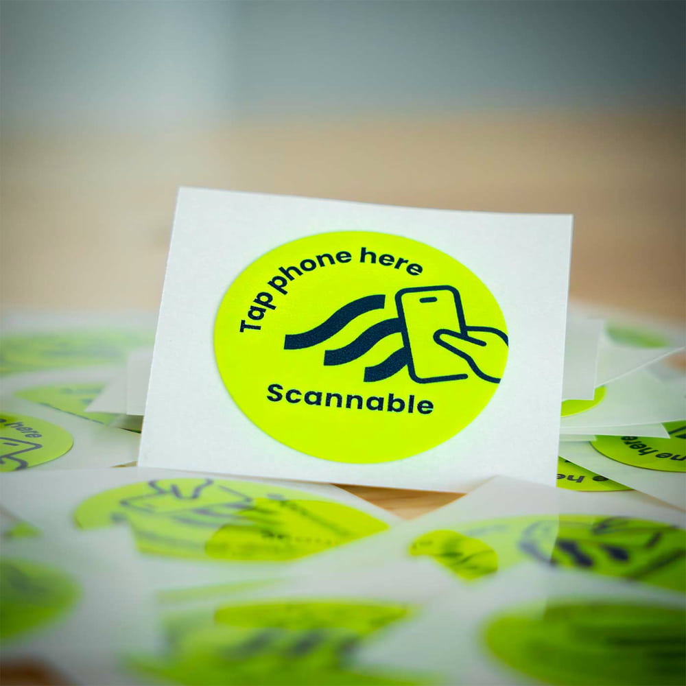 Gros plan d’un seul autocollant de casque rond jaune, autocollant NFC avec inscription noire « Tap phone here Scannable » et une icône de smartphone, entouré d’autres autocollants