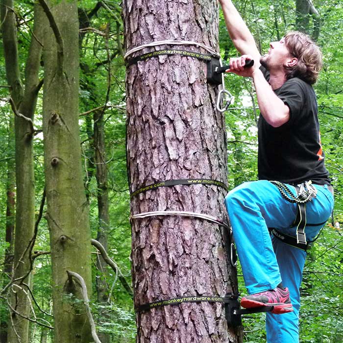 Tree-Monkey Steppers attaché au tronc d’arbre, homme avec jambe de pantalon bleu et t-shirt noir grimpe avec le pied sur le pied en métal et le harnais