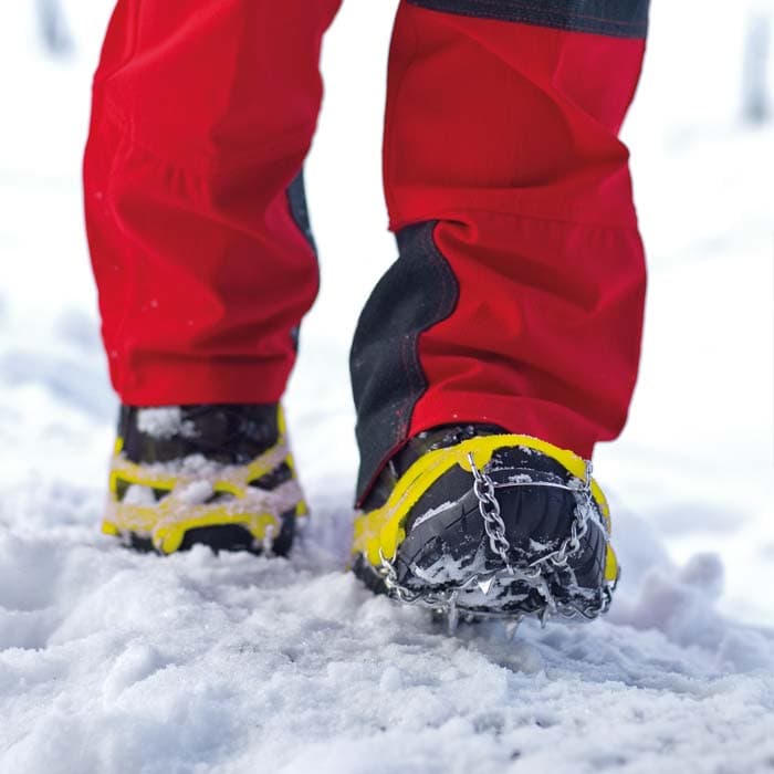 Pointes avec élastiques jaunes et chaînes métalliques sur des chaussures noires, portées par une personne en pantalon rouge dans la neige