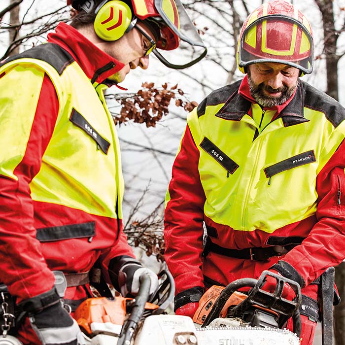 Deux personnes portent la veste forestière KlimaAIR® en jaune et rouge, une personne tient une tronçonneuse, toutes deux portent des casques de sécurité et des protections auditives.