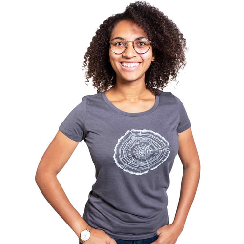 Une femme aux cheveux noirs bouclés et aux lunettes porte un t-shirt gris Treeslice Women avec un motif de tronc d’arbre blanc sur le devant