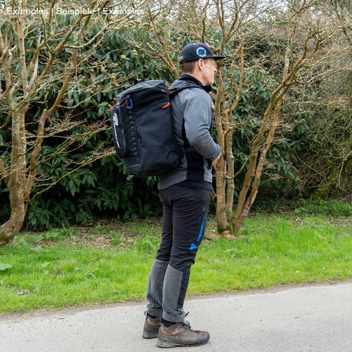 Personne avec un sac à dos noir bébé mule, fabricant non visible, debout sur le côté sur un chemin, entouré d’arbres et d’herbe