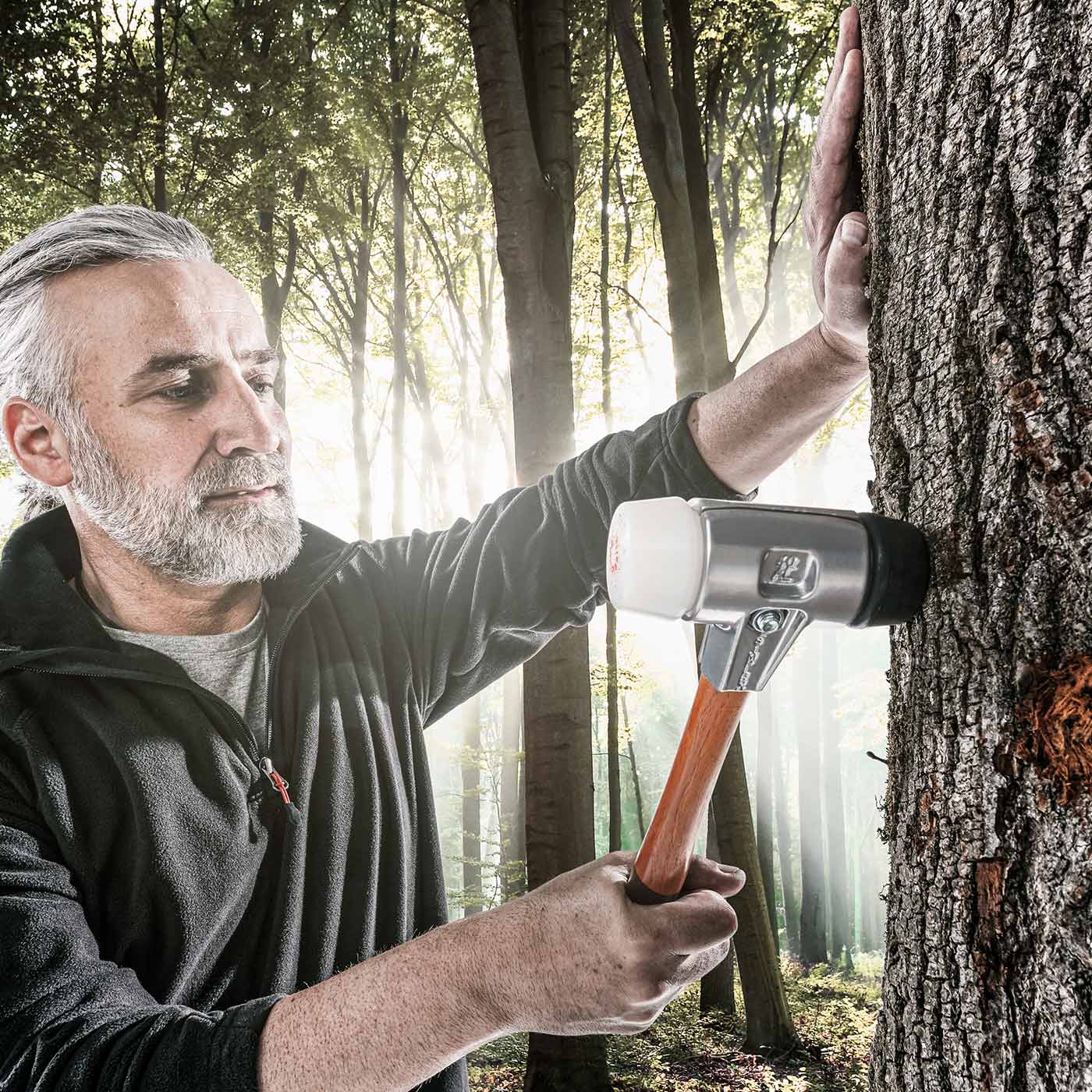 Un homme à la barbe grise et à la veste noire tient le Simplex Schönhammer 370 contre un tronc d’arbre dans la forêt
