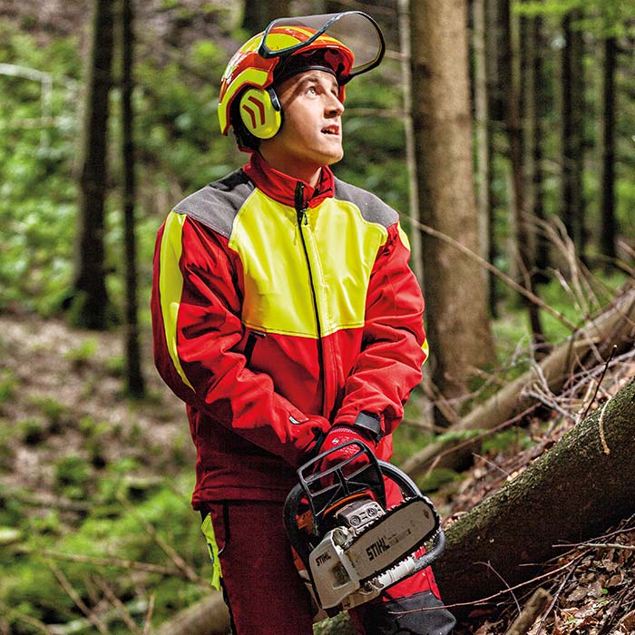 Homme portant une veste anti-coupure en rouge avec des accents jaunes et gris, casque et gants, tenant une tronçonneuse dans la forêt