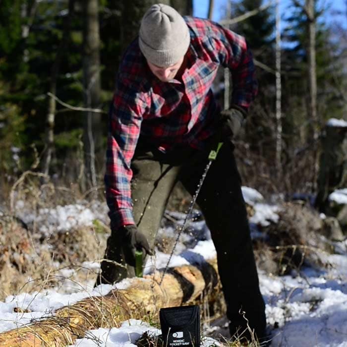 Personne avec une casquette grise et une chemise à carreaux utilisant la scie de poche nordique sur un tronc d’arbre, sac noir avec des lettres blanches couché sur le sol