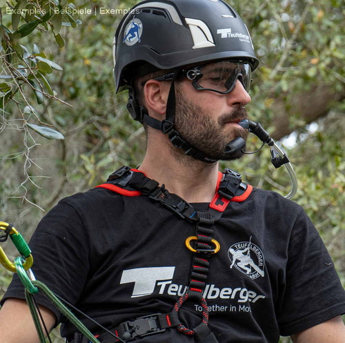 Homme portant un harnais d’escalade upMOTION SRT, un casque noir et des lunettes, tenant un tube d’abreuvement dans sa bouche, des arbres et des feuilles en arrière-plan