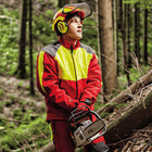 Homme portant une veste anti-coupure en rouge avec des accents jaunes et gris, casque et gants, tenant une tronçonneuse dans la forêt