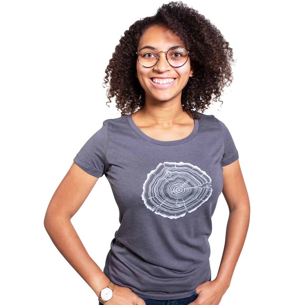 Une femme aux cheveux noirs bouclés et aux lunettes porte un t-shirt gris Treeslice Women avec un motif de tronc d’arbre blanc sur le devant