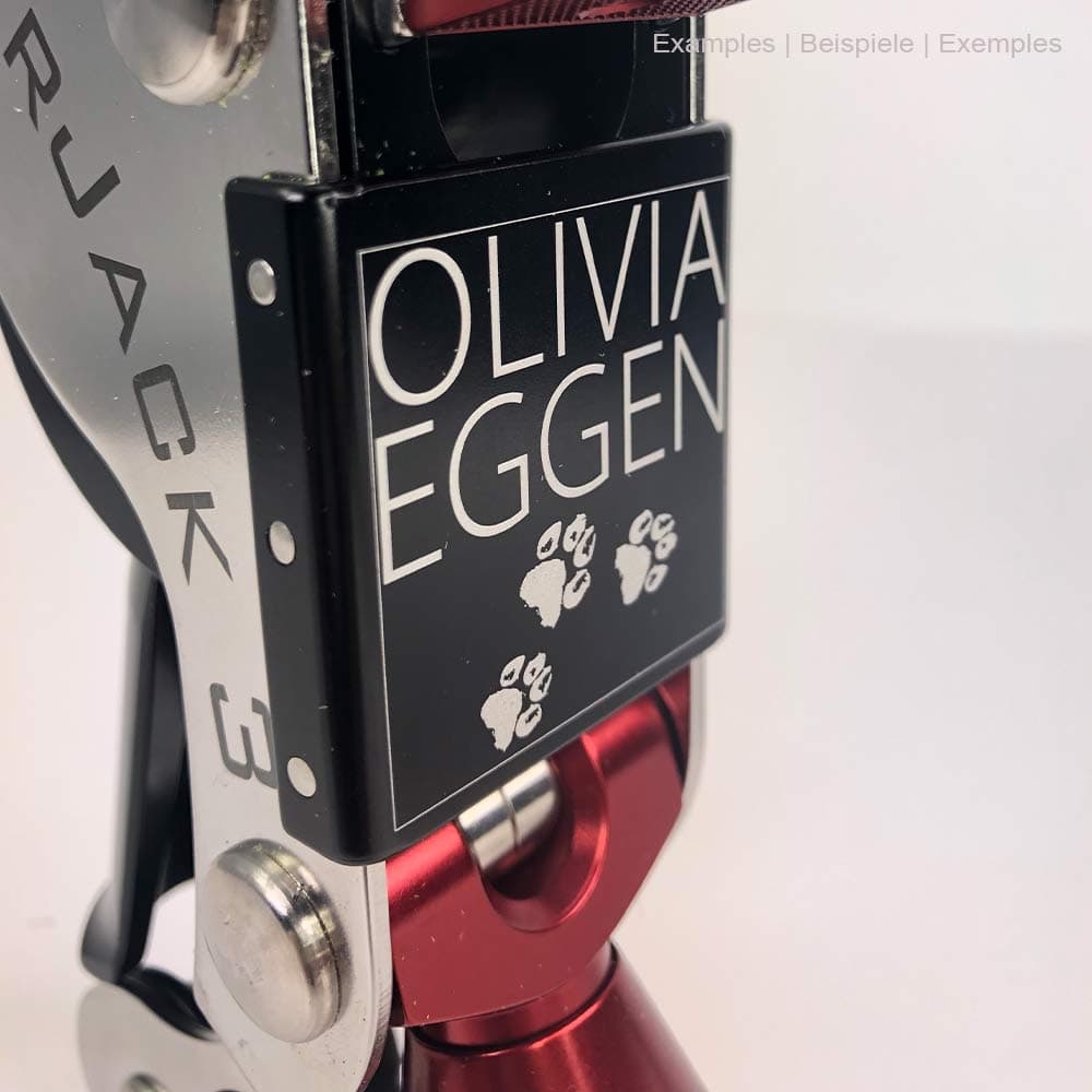 Gros plan du bouclier noir de la SpiderJack 3 avec gravure blanche « OLIVIA EGGEN » et empreintes de pattes, crochet en métal rouge et cadre argenté