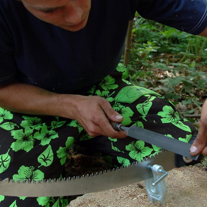 Personne en pantalon vert et noir tenant une lime grise et travaillant sur une scie à l’extérieur