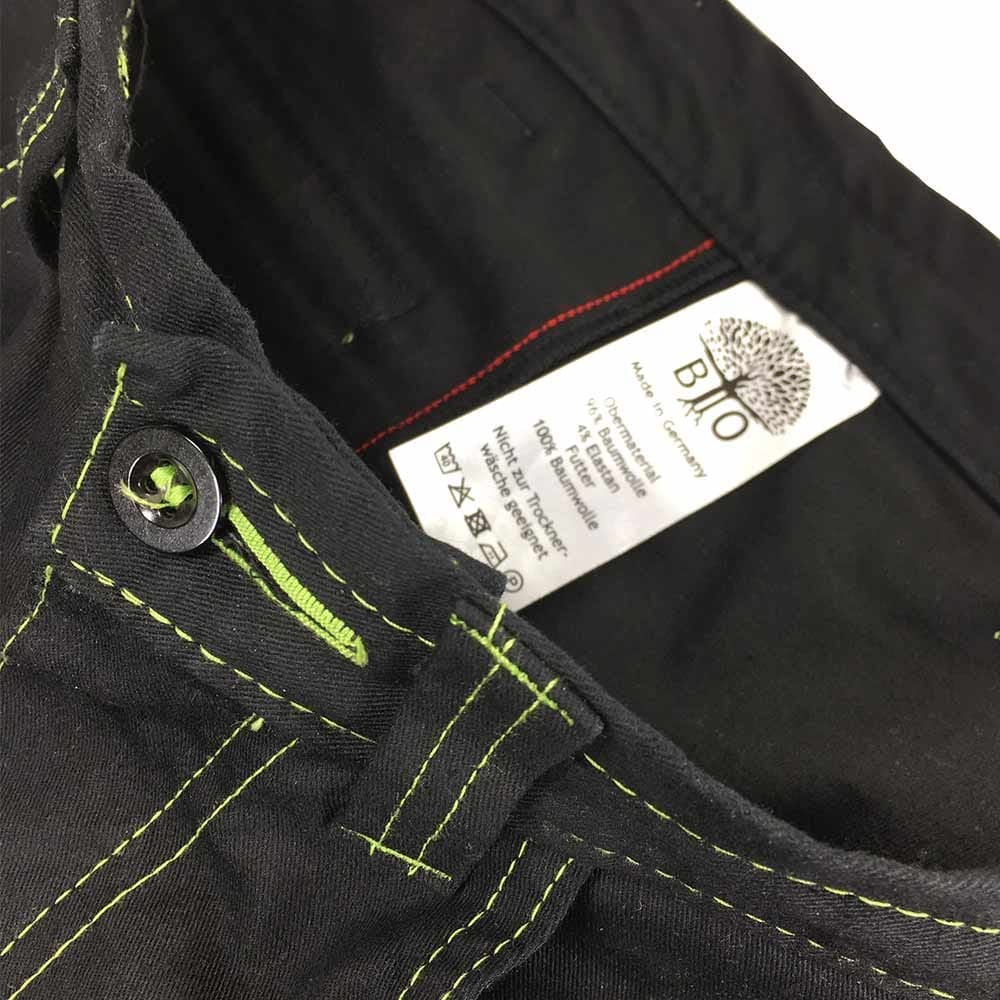Gros plan de la ceinture du pantalon d’extérieur Roots Pure en noir avec boutonnière verte et fil vert, à l’intérieur d’une étiquette blanche avec texte et logo de l’arbre