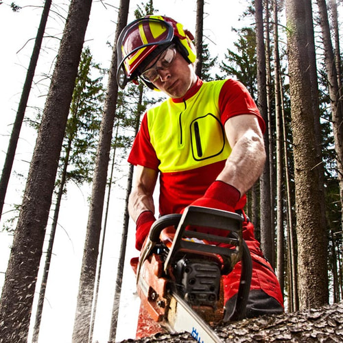 Homme portant la chemise à col zippé en rouge et jaune, casque de sécurité, lunettes et gants, tenant une tronçonneuse dans la forêt