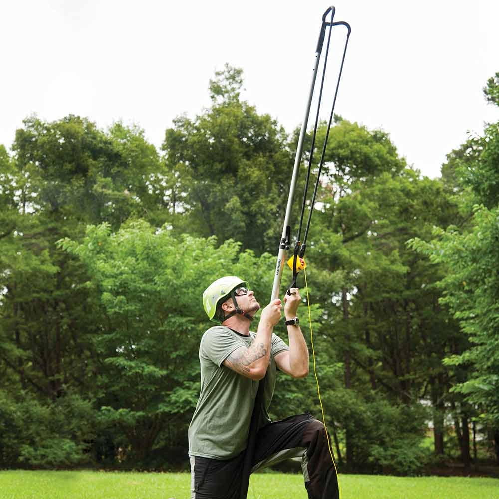 Personne en casque vert et lunettes agenouillée sur l’herbe verte et tenant une baguette Big Shot avec poignée noire et sangle jaune devant une forêt