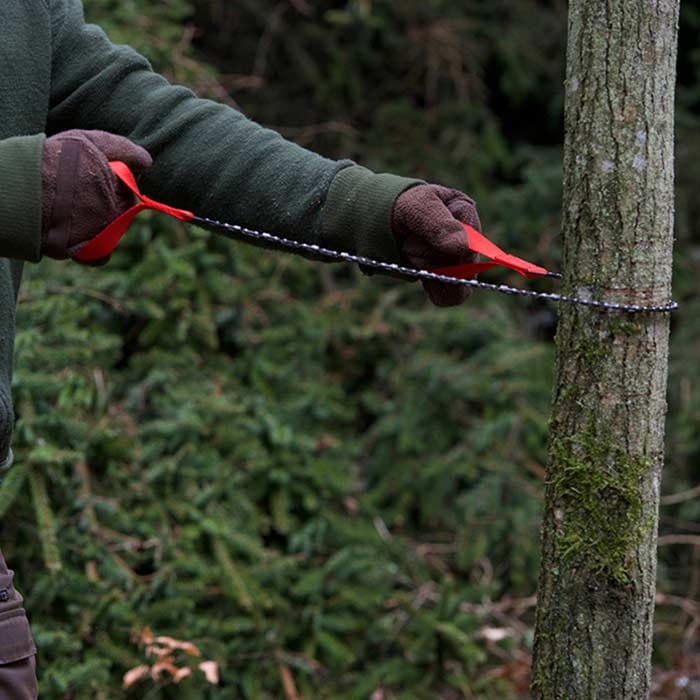 Personne en veste verte et gants tenant la scie de poche nordique avec des boucles rouges tendues autour d’un tronc d’arbre