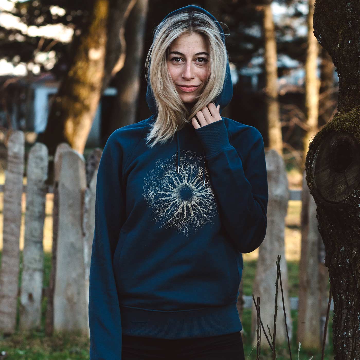 Une femme se tient à l’extérieur à côté d’un arbre, portant un sweat à capuche bleu en bois Iris avec une capuche et un motif blanc ressemblant à une racine d’arbre sur la poitrine.