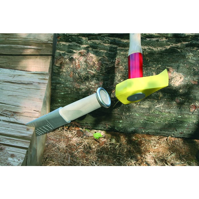 Alu 360, outil avec lame en aluminium et manche en bois clair avec finition métallique, à côté un marteau avec un manche rouge et un capuchon de protection jaune, tous deux posés sur du bois et de la terre