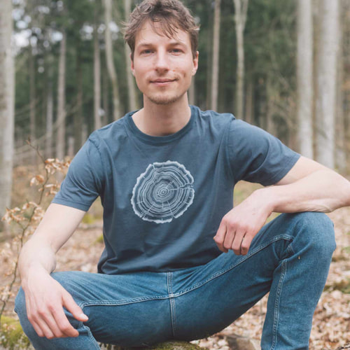 Un homme est assis dans la forêt et porte un T-shirt bleu foncé avec un tronc d’arbre blanc coupé, nom du produit Treeslice