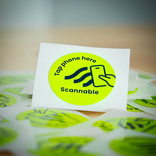 Gros plan d’un seul autocollant de casque rond jaune, autocollant NFC avec inscription noire « Tap phone here Scannable » et une icône de smartphone, entouré d’autres autocollants