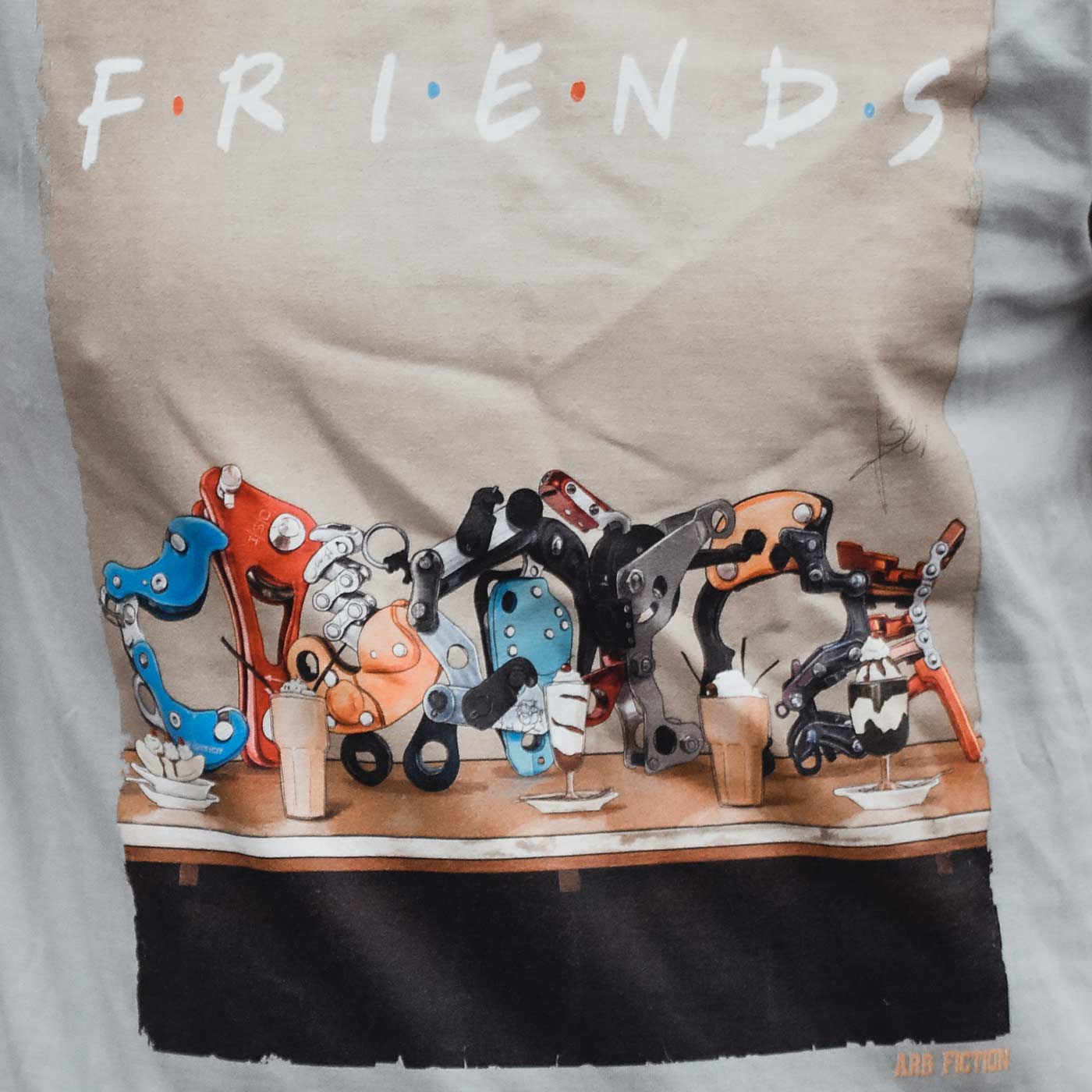 Gros plan de l’imprimé du T-shirt avec les mots « Friends » et des pièces mécaniques détaillées de différentes couleurs sur une table, avec le lettrage « ARB FICTION » visible en bas à droite