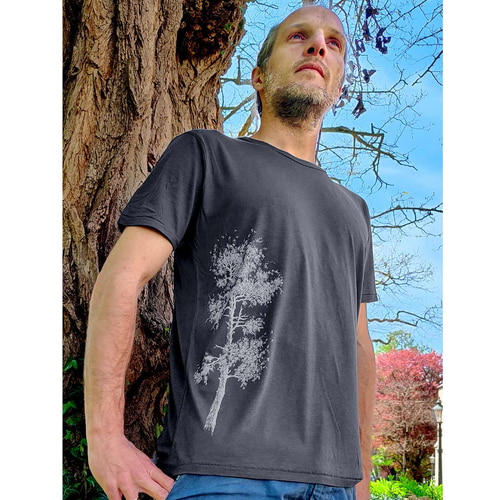Homme debout à l’extérieur devant un arbre portant un t-shirt gris foncé en forme de pin avec un imprimé d’arbre blanc sur le côté gauche de la chemise.