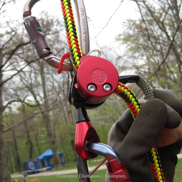 Aire de jeux LockJack 2 avec boîtier en métal rouge, mousqueton gris et corde jaune-rouge, tenue d’une main avec un gant gris sur fond de forêt