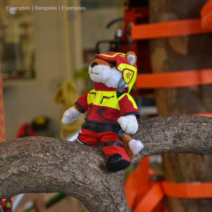 Figurine en peluche Wiffzack assise sur un tronc d’arbre, portant des vêtements rouges et jaunes et un casque, en arrière-plan l’environnement de l’atelier visible