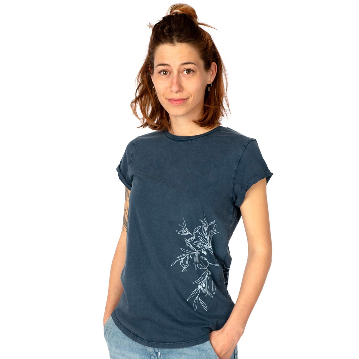 Une femme porte un T-shirt bleu foncé « Rameau d’olivier » avec un motif de branche d’olivier blanc sur le côté et a les mains dans les poches de son pantalon.