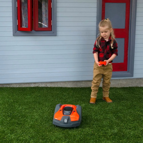 Un enfant vêtu d’une chemise à carreaux rouges, d’un pantalon beige et de bottes marron contrôle l’Automower orange et noir pour enfants de Husqvarna sur une pelouse verte devant une maison avec une fenêtre et un cadre de porte rouges