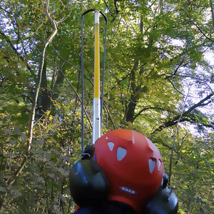 Personne avec un casque rouge et une protection auditive par derrière, tenant la tige Big Shot avec poignée noire et sangle jaune dans une forêt