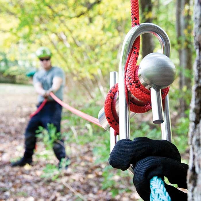 Port A Wrap Medium, appareil en métal argenté avec une corde rouge attachée à un arbre, en arrière-plan une personne floue avec un casque et des gants tirant sur une corde