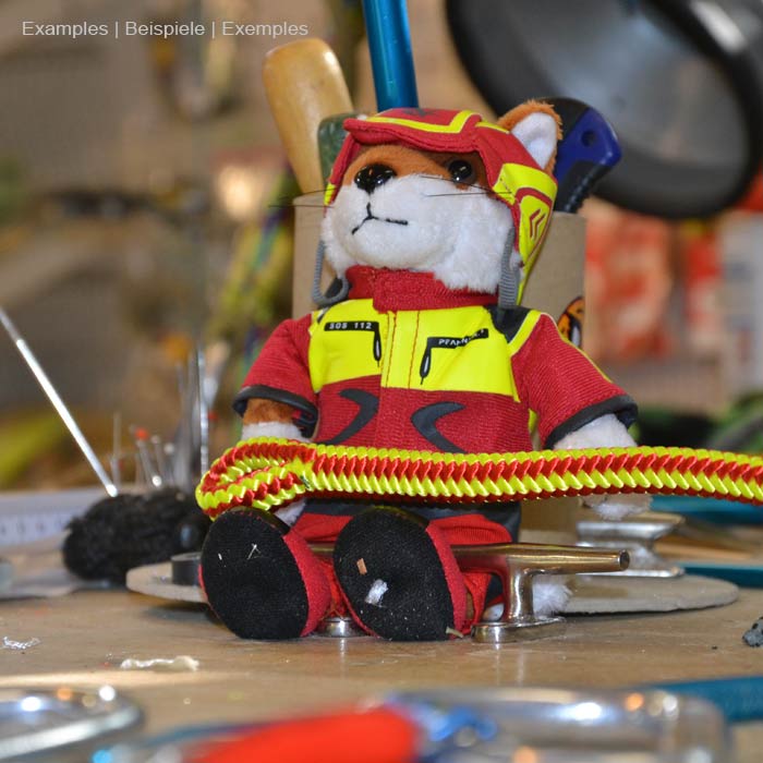 Figurine en peluche Wiffzack assise sur une table entourée d’outils, portant des vêtements rouges et jaunes et un casque