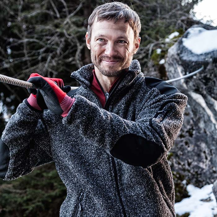Homme portant une veste grizzly, un cardigan gris avec une bordure noire sur les épaules et les coudes, un maillot de corps rouge et des gants rouges, une forêt et des rochers avec de la neige en arrière-plan